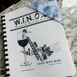 Puede incluir: Un libro de cocina encuadernado en espiral titulado "W.I.N.O. (Women in Need of Sanity) Cook with Wine" de Bonnie Jesseph. Un marcap&aacute;ginas de esquina con estampado azul y blanco con personajes de dibujos animados descansa sobre las p&aacute;ginas del libro.