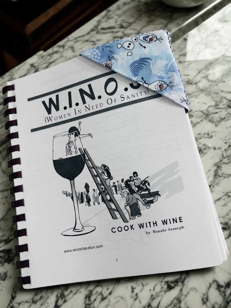 Puede incluir: Un libro de cocina encuadernado en espiral titulado "W.I.N.O. (Women in Need of Sanity) Cook with Wine" de Bonnie Jesseph. Un marcap&aacute;ginas de esquina con estampado azul y blanco con personajes de dibujos animados descansa sobre las p&aacute;ginas del libro.