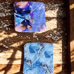 Puede incluir: Dos art&iacute;culos de tela doblados con dise&ntilde;os de Frozen. Uno presenta a Elsa y Anna en tonos azules, morados y rosas. El otro tiene un fondo azul claro con Olaf el mu&ntilde;eco de nieve. Ambos est&aacute;n sobre una superficie de madera.