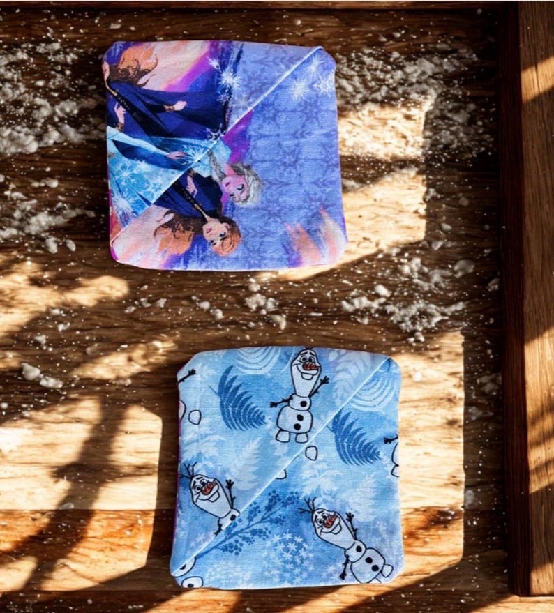 Puede incluir: Dos art&iacute;culos de tela doblados con dise&ntilde;os de Frozen. Uno presenta a Elsa y Anna en tonos azules, morados y rosas. El otro tiene un fondo azul claro con Olaf el mu&ntilde;eco de nieve. Ambos est&aacute;n sobre una superficie de madera.