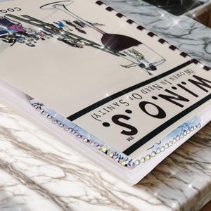 Puede incluir: Un libro de cocina titulado "W.I.N.O.S. (Women In Need Of Sanity)" con una ilustraci&oacute;n de copa de vino. La portada es de color blanco roto con texto negro y un borde decorativo. El libro est&aacute; sobre una superficie de m&aacute;rmol.