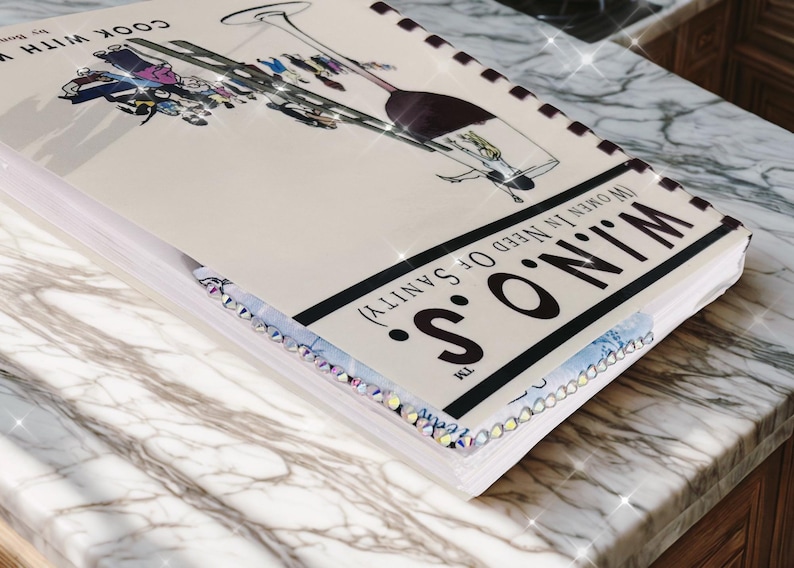 Puede incluir: Un libro de cocina titulado "W.I.N.O.S. (Women In Need Of Sanity)" con una ilustraci&oacute;n de copa de vino. La portada es de color blanco roto con texto negro y un borde decorativo. El libro est&aacute; sobre una superficie de m&aacute;rmol.
