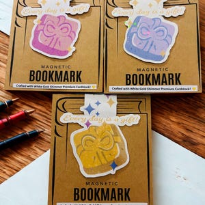 May include: Three magnetic bookmarks with gift box designs in purple, blue, and yellow. Each bookmark has the text "Every day is a gift!" and "MAGNETIC BOOKMARK." The bookmarks are crafted with white gold shimmer premium cardstock.