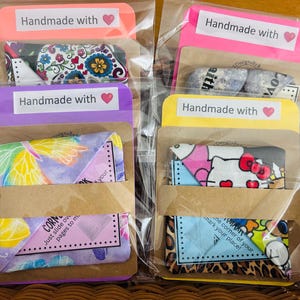 Puede incluir: Varios marcadores de tela en envases transparentes, cada uno con un dise&ntilde;o diferente. Los marcadores est&aacute;n empaquetados en una funda de papel marr&oacute;n y est&aacute;n etiquetados "Handmade with" con un coraz&oacute;n. Los dise&ntilde;os incluyen patrones florales, de mariposas y de Hello Kitty.