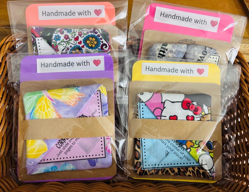 Puede incluir: Varios marcadores de tela en envases transparentes, cada uno con un dise&ntilde;o diferente. Los marcadores est&aacute;n empaquetados en una funda de papel marr&oacute;n y est&aacute;n etiquetados "Handmade with" con un coraz&oacute;n. Los dise&ntilde;os incluyen patrones florales, de mariposas y de Hello Kitty.
