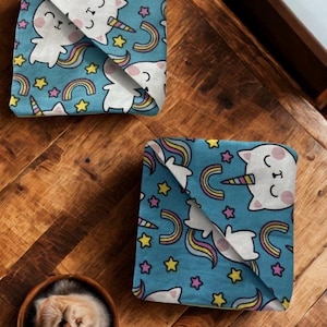 Puede incluir: Dos cuadrados de tela doblados con un fondo azul con gatos-unicornios blancos, arco&iacute;ris y estrellas. Los art&iacute;culos est&aacute;n sobre una superficie de madera. Un peque&ntilde;o cuenco redondo de madera con un gato marr&oacute;n claro est&aacute; en la esquina inferior izquierda.