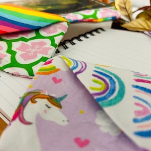 Puede incluir: Primer plano de varios art&iacute;culos de tela con dise&ntilde;os coloridos. Uno presenta un dise&ntilde;o de arco&iacute;ris, otro flores rosas sobre un fondo verde y un tercero un unicornio con una melena de arco&iacute;ris. Un cuaderno de espiral est&aacute; en el fondo.