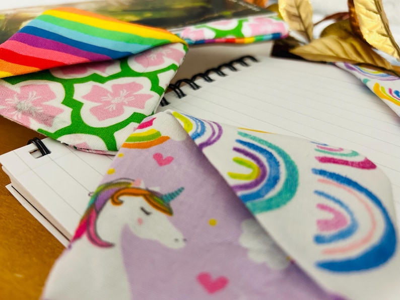 Puede incluir: Primer plano de varios art&iacute;culos de tela con dise&ntilde;os coloridos. Uno presenta un dise&ntilde;o de arco&iacute;ris, otro flores rosas sobre un fondo verde y un tercero un unicornio con una melena de arco&iacute;ris. Un cuaderno de espiral est&aacute; en el fondo.