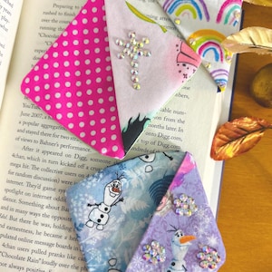 Puede incluir: Marcadores de libros de tela hechos a mano. Un marcador presenta un patr&oacute;n de lunares rosas, otro un dise&ntilde;o de arco&iacute;ris, y otros dos muestran al personaje Olaf de Frozen. Cada marcador est&aacute; adornado con brillantes pedrer&iacute;a.