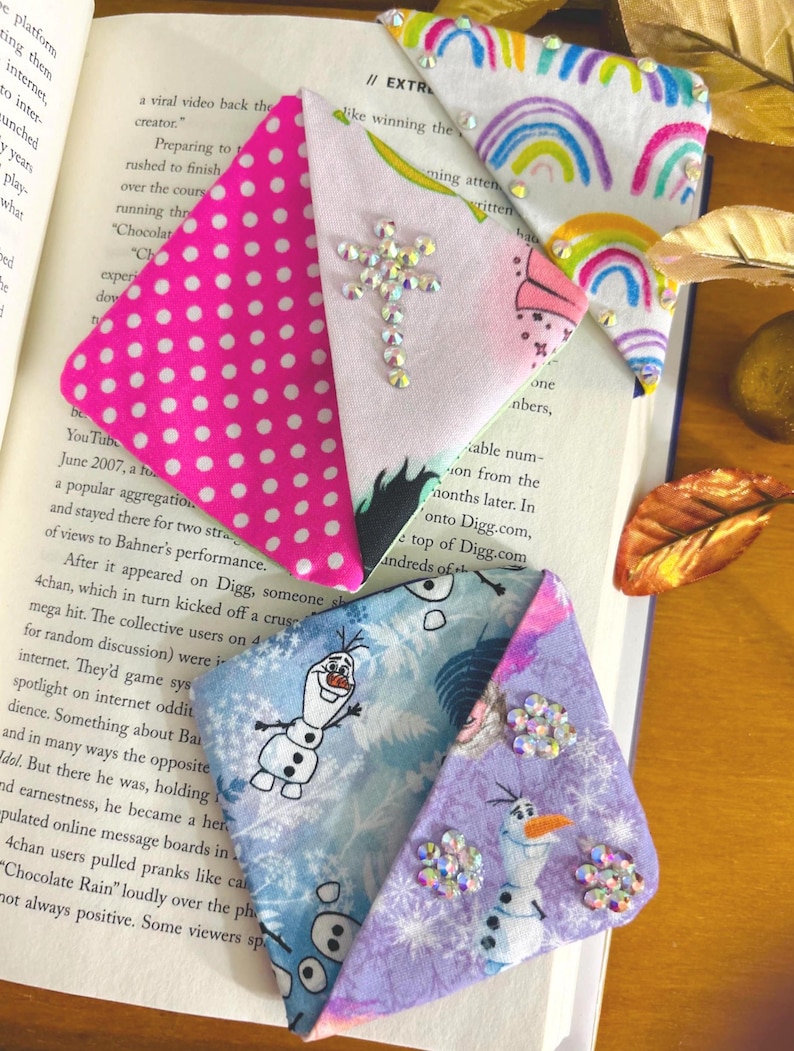 Puede incluir: Marcadores de libros de tela hechos a mano. Un marcador presenta un patr&oacute;n de lunares rosas, otro un dise&ntilde;o de arco&iacute;ris, y otros dos muestran al personaje Olaf de Frozen. Cada marcador est&aacute; adornado con brillantes pedrer&iacute;a.