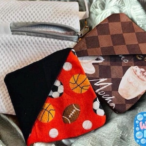 Puede incluir: Una bolsa de lavander&iacute;a de malla blanca con cremallera y dos peque&ntilde;as bolsas triangulares. Una bolsa es negra y roja con dise&ntilde;os de balones deportivos. La otra es marr&oacute;n con un dise&ntilde;o de caf&eacute;. Incluye el texto "Wash Me!"