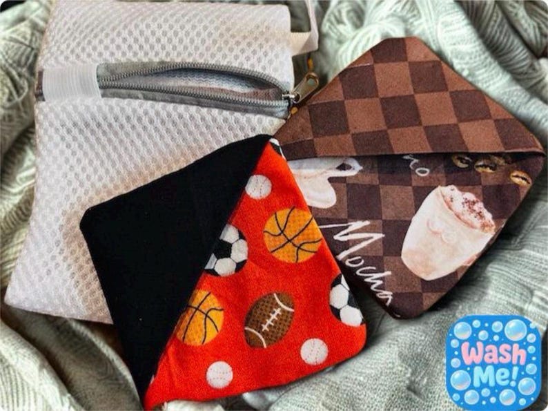 Puede incluir: Una bolsa de lavander&iacute;a de malla blanca con cremallera y dos peque&ntilde;as bolsas triangulares. Una bolsa es negra y roja con dise&ntilde;os de balones deportivos. La otra es marr&oacute;n con un dise&ntilde;o de caf&eacute;. Incluye el texto "Wash Me!"