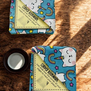 Puede incluir: Dos marcadores de esquina con un dise&ntilde;o caprichoso. Los marcadores presentan un fondo azul claro con gatos-unicornios blancos, arco&iacute;ris y estrellas. Cada marcador tiene una esquina amarilla con el texto "CORNER BOOKMARK. Just slide over the corner of your pages to mark your place!"