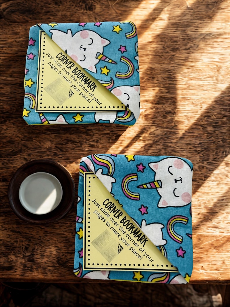 Puede incluir: Dos marcadores de esquina con un dise&ntilde;o caprichoso. Los marcadores presentan un fondo azul claro con gatos-unicornios blancos, arco&iacute;ris y estrellas. Cada marcador tiene una esquina amarilla con el texto "CORNER BOOKMARK. Just slide over the corner of your pages to mark your place!"