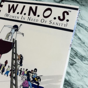 Puede incluir: Un libro de cocina titulado "W.I.N.O.S. (Women In Need Of Sanity)" con una ilustraci&oacute;n de dibujos animados de personas subiendo a una copa de vino. La portada es de color blanquecino con texto negro y burdeos y un borde azul oscuro. Las palabras "COOK WITH" son visibles.