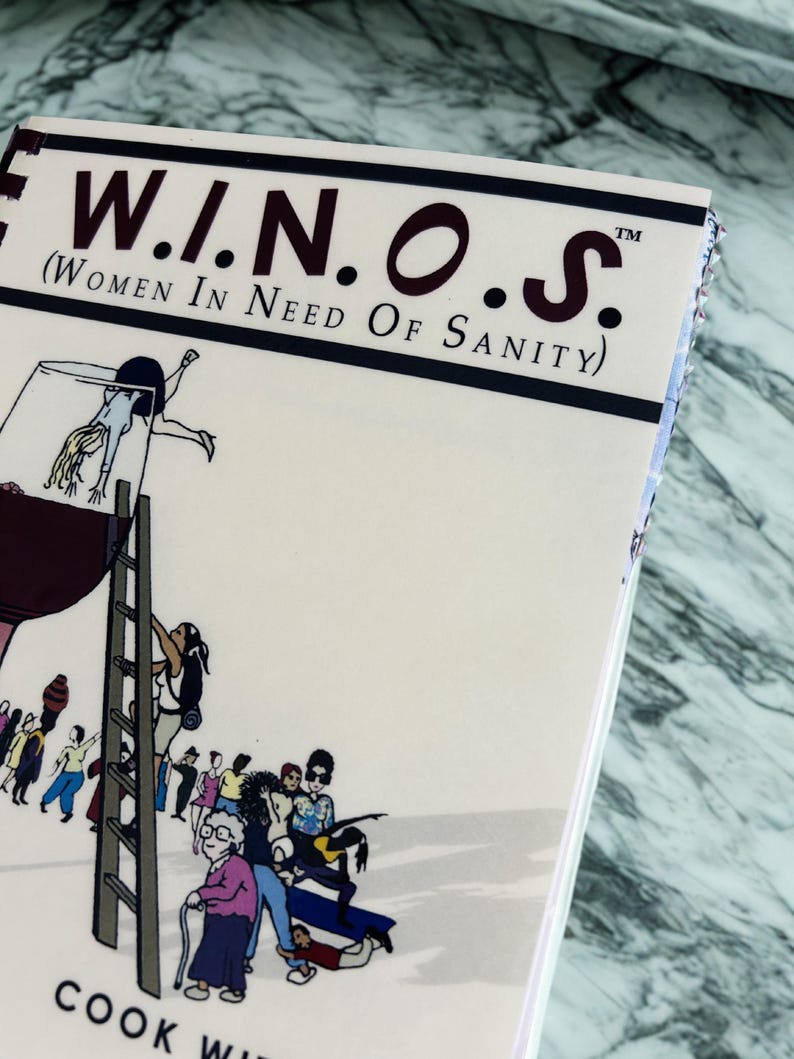 Puede incluir: Un libro de cocina titulado "W.I.N.O.S. (Women In Need Of Sanity)" con una ilustraci&oacute;n de dibujos animados de personas subiendo a una copa de vino. La portada es de color blanquecino con texto negro y burdeos y un borde azul oscuro. Las palabras "COOK WITH" son visibles.