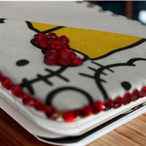 Puede incluir: Un cuaderno blanco con un dise&ntilde;o de Hello Kitty, con contornos negros y una secci&oacute;n amarilla. El cuaderno est&aacute; bordeado con joyas rojas. El dise&ntilde;o incluye la cara de Hello Kitty y un lazo rojo.