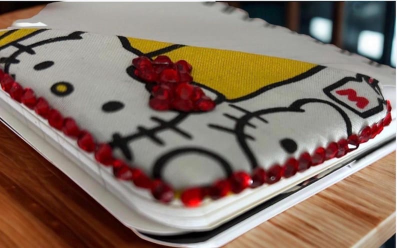 Puede incluir: Un cuaderno blanco con un dise&ntilde;o de Hello Kitty, con contornos negros y una secci&oacute;n amarilla. El cuaderno est&aacute; bordeado con joyas rojas. El dise&ntilde;o incluye la cara de Hello Kitty y un lazo rojo.