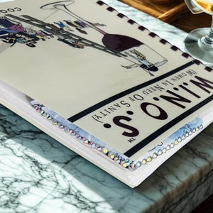 Puede incluir: Un libro de cocina titulado "Cook With W.I.N.O.S. (Women in Need of Sanity)" con una ilustraci&oacute;n de copa de vino. El libro tiene un borde decorativo y est&aacute; sobre una superficie de m&aacute;rmol.