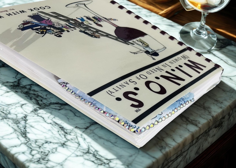 Puede incluir: Un libro de cocina titulado "Cook With W.I.N.O.S. (Women in Need of Sanity)" con una ilustraci&oacute;n de copa de vino. El libro tiene un borde decorativo y est&aacute; sobre una superficie de m&aacute;rmol.