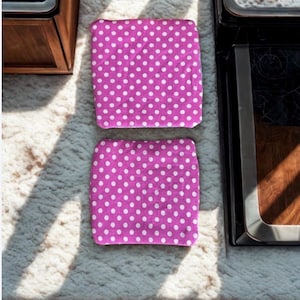 Puede incluir: Dos art&iacute;culos cuadrados de tela magenta con un patr&oacute;n de lunares blancos. Se colocan sobre una superficie clara y texturizada. Un mueble de madera y un electrodom&eacute;stico negro son visibles en el fondo.