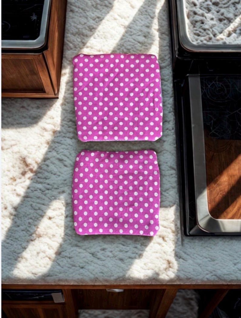 Puede incluir: Dos art&iacute;culos cuadrados de tela magenta con un patr&oacute;n de lunares blancos. Se colocan sobre una superficie clara y texturizada. Un mueble de madera y un electrodom&eacute;stico negro son visibles en el fondo.