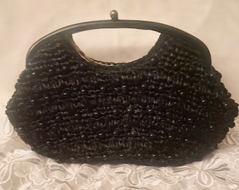 May include: A black woven clutch bag with a black top handle. The bag is made of a textured material and has a rounded shape.