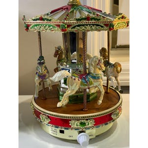 May include: A decorative carousel with several toy horses. The carousel has a red, green, and gold canopy and a wooden base. The horses are cream-colored with colorful saddles. The carousel has a power switch and a power input.