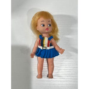 May include: A vintage doll with blonde hair and blue eyes. The doll wears a blue skirt and a striped top with red, white, yellow, and black accents. A white belt with a silver buckle cinches the waist. The doll is standing.