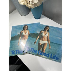 May include: Two vinyl records featuring a woman in a white bikini standing in the ocean. The records have a blue and white color scheme with the text "To The Good Life" in blue.