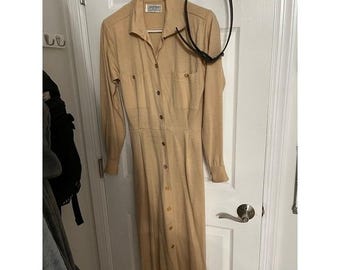 May include: A tan-colored, long-sleeved dress with a collared neckline and button-down front. The dress features two chest pockets and a fitted waist. A black belt hangs on the door behind the dress.
