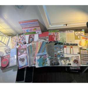 May include: A collection of scrapbooking and crafting supplies, including patterned paper, stickers, stamps, and embellishments. The items are packaged and arranged on a white surface, with a variety of colors and designs visible. The assortment includes alphabet stickers and decorative tape.