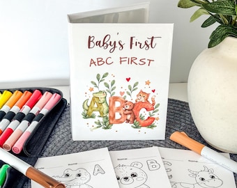 May include: A white booklet titled Baby's First ABC FIRST featuring a watercolor illustration of a crocodile, bear, and cat with the letters A, B, and C. Below are coloring pages of a crocodile, bear, and cow with markers nearby.