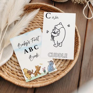 Può includere: Due libri per bambini in un cestino di vimini. Un libro è intitolato "Baby's First ABC Book" con illustrazioni dei personaggi di Winnie the Pooh. L'altra carta dice "C is for Cuddle" con un'illustrazione di Winnie the Pooh.