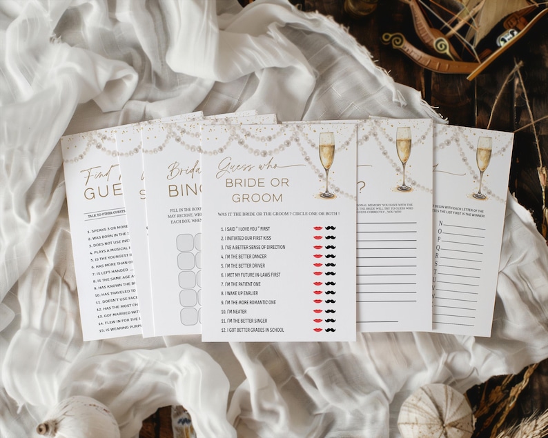 May include: A set of white bridal shower game cards with gold detailing and champagne glass illustrations. The cards feature games such as "Find the Guest", "Bride Bingo", and "Guess Who: Bride or Groom?" The cards are displayed on a white fabric background.