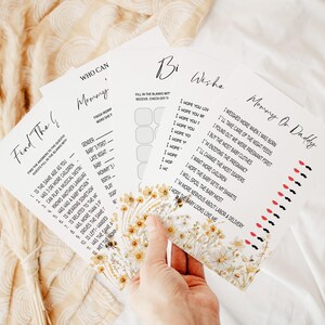 May include: A set of four baby shower game cards with floral accents. The cards include prompts like "Who Can Mommy", "Baby Wishes", and "Mommy or Daddy". The cards are held in a hand.