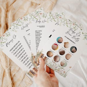 May include: A collection of baby shower game cards with floral and botanical designs. The cards feature text such as "Mommy Or," "Baby Or Beer Belly," and "Would Who?" The cards are held by a person.
