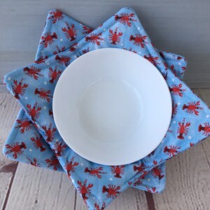 May include: A white ceramic bowl rests on two blue fabric bowl cozies. The cozies are patterned with red lobsters and white polka dots. The bowl cozies are square and folded to hold the bowl.