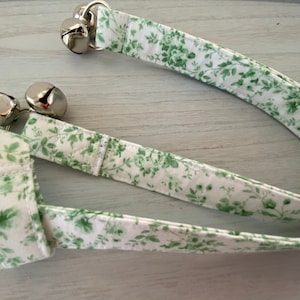 May include: A white and green floral patterned collar with two silver bells. The collar is made of fabric and has a stitched design. The bells are attached with silver metal rings. The overall theme is a pet accessory.