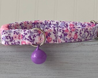 May include: A purple and pink floral patterned cat collar with a purple bell. The collar has a pink buckle and a silver ring.