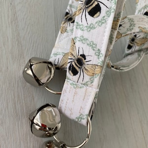 May include: A white fabric dog collar with a bee pattern and three silver bells attached.