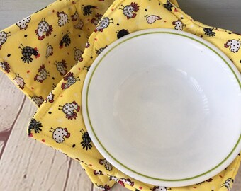 May include: Two yellow fabric bowl cozies with a black and white chicken pattern. The cozies are folded and a white bowl is sitting inside.