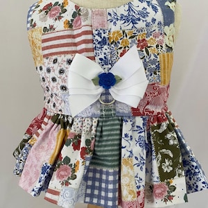 May include: A sleeveless harness dress featuring a multi-colored patchwork pattern of floral, striped, and checkered fabrics in shades of blue, pink, yellow, and green. A large white ribbon bow with a small blue fabric rose sits at the center chest.