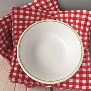May include: A white ceramic bowl with a green rim sits atop a red and white checkered bowl cozy. The cozy is square-shaped and quilted, providing a heat-resistant surface for serving hot dishes. The image is a close-up shot.