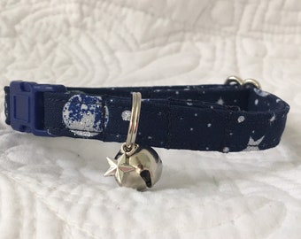 Pode incluir: Um colar de gato azul marinho com um padrão de lua e estrelas brancas. O colar tem um sino e um amuleto de estrela prateados.