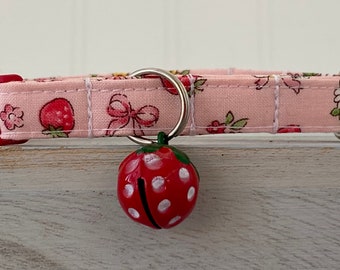 May include: Pink cat collar with a strawberry print and a red strawberry-shaped bell.