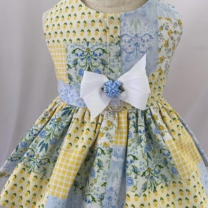 May include: A sleeveless pet harness dress featuring a patchwork pattern of yellow and blue floral prints, yellow gingham, and blue damask motifs. The bodice is accented with a white ribbon bow and a small blue fabric rose above a metal D-ring.