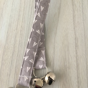 May include: A gray fabric strap with a white paper airplane pattern and three silver jingle bells attached.