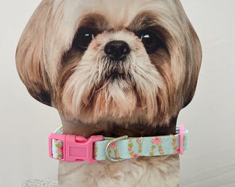 May include: A dog collar with a light blue background and a pink buckle. The collar has a floral pattern with pink roses and green leaves. The collar is displayed on a wooden stand with a dog's face.