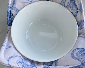 May include: A white ceramic bowl rests on a square, quilted pot holder. The pot holder features a blue and white toile pattern with rooster and floral designs. The bowl is centered on the pot holder.